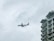 An airplane is in flight against a cloudy sky, with a multi-story building on the right side of the image. The building has balconies and a distinct architectural design on the roof.