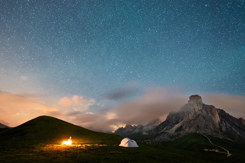 Tienda blanca en campo de hierba verde cerca de la montaña bajo el cielo azul con estrellas durante la noche