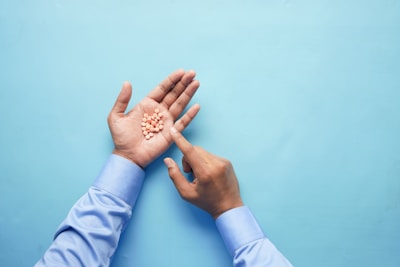 A pair of hands with light skin, one of which is holding a pile of small orange pills against a light blue background. The person is wearing a light blue shirt, and one hand is pointing to the pills.