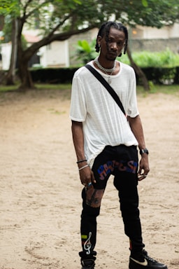 A person stands in a casual outdoor setting, wearing a loose white T-shirt and black pants with colorful designs. The individual has braided hair and accessorizes with a silver necklace and bracelets. The background includes trees and greenery with a hint of urban structures.
