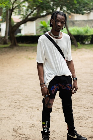 A person stands in a casual outdoor setting, wearing a loose white T-shirt and black pants with colorful designs. The individual has braided hair and accessorizes with a silver necklace and bracelets. The background includes trees and greenery with a hint of urban structures.