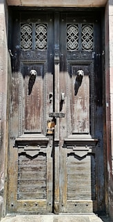 An old, weathered wooden door with intricate wrought iron designs above two lion head door knockers. The wood appears aged with visible peeling paint and a rustic texture. The door is locked with a sliding bolt and a padlock.