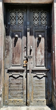 An old, weathered wooden door with intricate wrought iron designs above two lion head door knockers. The wood appears aged with visible peeling paint and a rustic texture. The door is locked with a sliding bolt and a padlock.