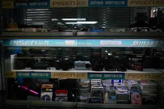 A display case with shelves containing various photography equipment and accessories, including cameras, lenses, cables, and film. The display is illuminated by lights and features branding for PISEN.