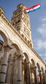 A tall stone bell tower with intricate carvings and arches rises majestically against a blue sky. The Croatian flag with red, white, and blue stripes waves prominently at the top, adding a touch of national pride.