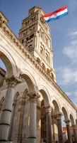 A tall stone bell tower with intricate carvings and arches rises majestically against a blue sky. The Croatian flag with red, white, and blue stripes waves prominently at the top, adding a touch of national pride.