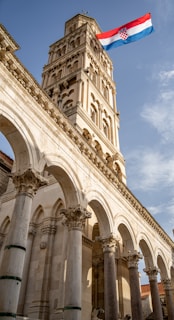 A tall stone bell tower with intricate carvings and arches rises majestically against a blue sky. The Croatian flag with red, white, and blue stripes waves prominently at the top, adding a touch of national pride.