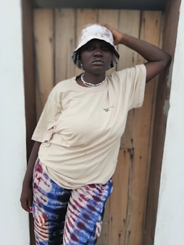 A person wearing a beige t-shirt, patterned pants, and a bucket hat poses casually in front of a wooden backdrop. They lean against the wall with one arm raised, expressing a relaxed demeanor. A beaded necklace adds detail to the outfit.
