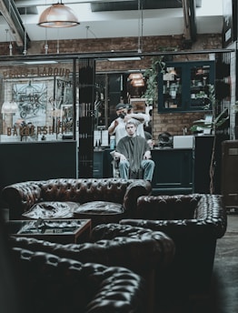 A modern, rustic barbershop interior with leather couches in the foreground. A person is receiving a haircut from a barber in the background, surrounded by industrial-style decor, exposed brick walls, and warm lighting fixtures.