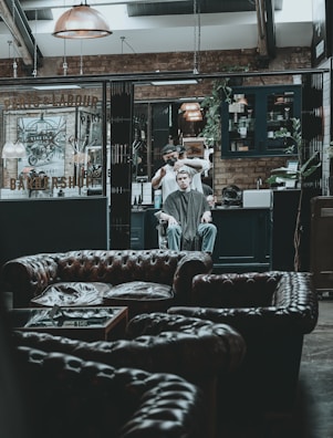 A modern, rustic barbershop interior with leather couches in the foreground. A person is receiving a haircut from a barber in the background, surrounded by industrial-style decor, exposed brick walls, and warm lighting fixtures.