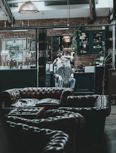 A modern, rustic barbershop interior with leather couches in the foreground. A person is receiving a haircut from a barber in the background, surrounded by industrial-style decor, exposed brick walls, and warm lighting fixtures.
