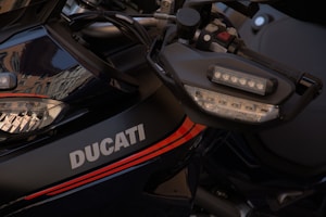 A close-up of a motorcycle featuring the brand name Ducati prominently displayed. The image captures part of the handlebar with a focus on the intricate design of the side mirror and front light. Reflections of buildings are visible on the glossy surface, adding an artistic element to the photograph.