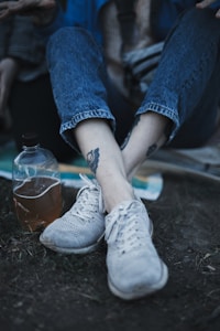 A person wearing blue jeans and white sneakers is sitting on the ground. They have tattoos on their ankles. Next to them is a clear plastic bottle filled with a brown liquid. The background includes a blurred blanket or cloth.