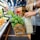 A person holding a shopping basket filled with fresh green basil, carrot tops, a paper bag, and a glass bottle of milk. The scene is set in a grocery store with fruits and vegetables displayed on shelves in the background.