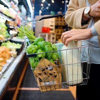 A person holding a shopping basket filled with fresh green basil, carrot tops, a paper bag, and a glass bottle of milk. The scene is set in a grocery store with fruits and vegetables displayed on shelves in the background.