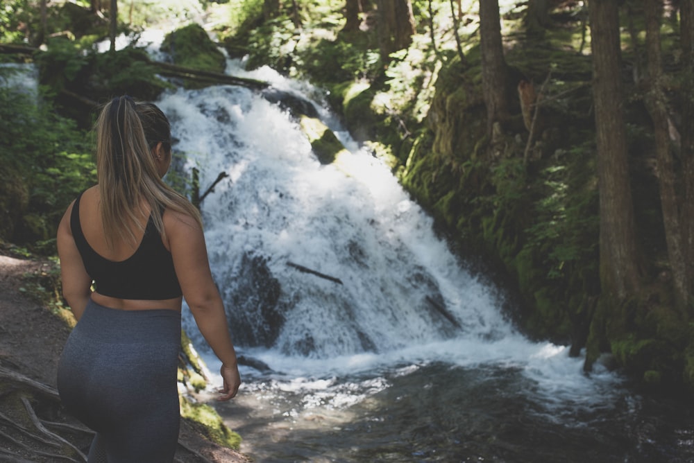woman in black tank top and blue denim jeans standing in front of waterfalls during daytime