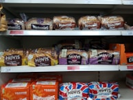 Several varieties of packaged bread are displayed on supermarket shelves. Brands such as Warburtons and Hovis are prominently visible, with loaves of wholemeal, seed sensations, and soft white varieties lined up in neat rows.