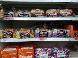 Several varieties of packaged bread are displayed on supermarket shelves. Brands such as Warburtons and Hovis are prominently visible, with loaves of wholemeal, seed sensations, and soft white varieties lined up in neat rows.