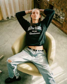 A person sits in a relaxed posture on a beige swivel chair. They are wearing a dark sweatshirt with 'BLUE ZONE' printed on it and light-colored ripped jeans. The setting appears to be indoors, with a light green carpet and wooden furniture in the background.