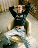A person sits in a relaxed posture on a beige swivel chair. They are wearing a dark sweatshirt with 'BLUE ZONE' printed on it and light-colored ripped jeans. The setting appears to be indoors, with a light green carpet and wooden furniture in the background.