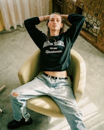 A person sits in a relaxed posture on a beige swivel chair. They are wearing a dark sweatshirt with 'BLUE ZONE' printed on it and light-colored ripped jeans. The setting appears to be indoors, with a light green carpet and wooden furniture in the background.