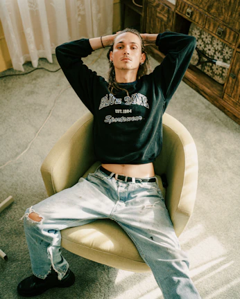 A person sits in a relaxed posture on a beige swivel chair. They are wearing a dark sweatshirt with 'BLUE ZONE' printed on it and light-colored ripped jeans. The setting appears to be indoors, with a light green carpet and wooden furniture in the background.