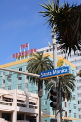 A vibrant cityscape featuring a building with a prominent red sign reading 'The Georgian' in bold letters, a tall palm tree, and a street sign for Santa Monica. The sky is clear with a few scattered clouds, and a traffic light hangs in the scene.