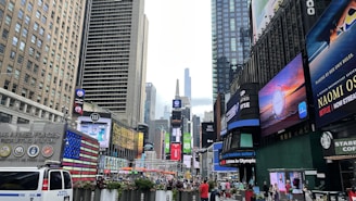 A bustling urban scene filled with tall buildings, bright digital billboards, and a crowd of people. Advertisements and logos are prominently displayed on the sides of the buildings. The street is busy with pedestrians and vehicles, including a visible NYPD police van. The vibrant energy of a city square is present, showcasing commercial activity and vibrant signage.