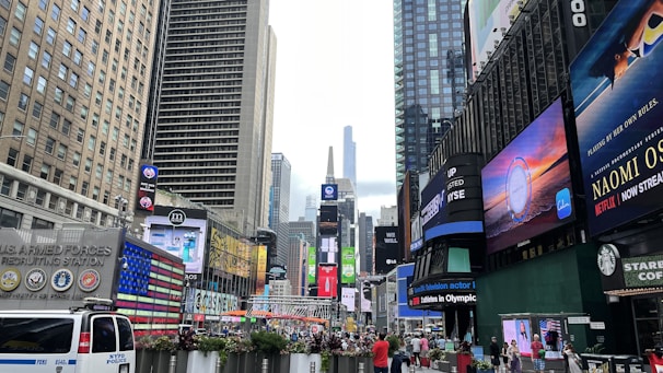 A bustling urban scene filled with tall buildings, bright digital billboards, and a crowd of people. Advertisements and logos are prominently displayed on the sides of the buildings. The street is busy with pedestrians and vehicles, including a visible NYPD police van. The vibrant energy of a city square is present, showcasing commercial activity and vibrant signage.