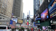 A bustling urban scene filled with tall buildings, bright digital billboards, and a crowd of people. Advertisements and logos are prominently displayed on the sides of the buildings. The street is busy with pedestrians and vehicles, including a visible NYPD police van. The vibrant energy of a city square is present, showcasing commercial activity and vibrant signage.