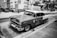 A vintage car is parked on a street with a sign on its roof advertising the sale and purchase of gold, silver, and watches. The surroundings include palm trees and a row of modern cars, set against high-rise buildings, reflecting a blend of classic and contemporary urban elements.