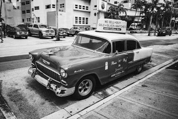 A vintage car is parked on a street with a sign on its roof advertising the sale and purchase of gold, silver, and watches. The surroundings include palm trees and a row of modern cars, set against high-rise buildings, reflecting a blend of classic and contemporary urban elements.