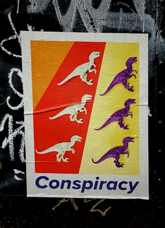 A colorful poster features illustrations of dinosaurs, with three white dinosaurs on a red background and three purple dinosaurs on a yellow background. The word 'Conspiracy' is written in bold blue letters at the bottom. The background behind the poster has a rough, black graffiti-covered surface.
