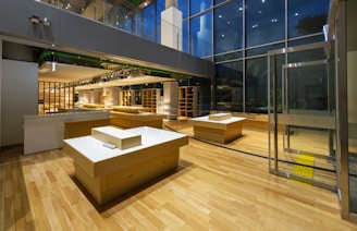 A modern, empty retail space with wooden flooring and large glass windows. Several wooden display tables are strategically placed throughout the room. The lighting is warm and inviting, highlighting the contemporary design elements of the space.
