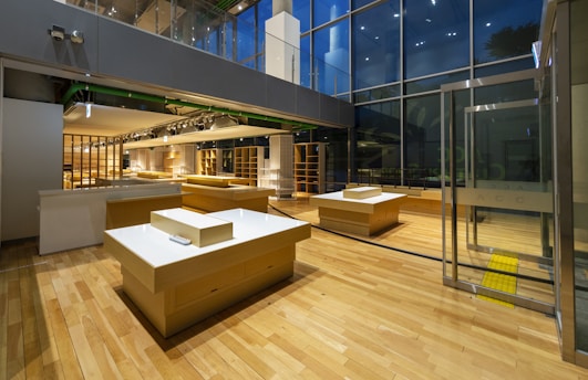 A modern, empty retail space with wooden flooring and large glass windows. Several wooden display tables are strategically placed throughout the room. The lighting is warm and inviting, highlighting the contemporary design elements of the space.