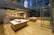 A modern, empty retail space with wooden flooring and large glass windows. Several wooden display tables are strategically placed throughout the room. The lighting is warm and inviting, highlighting the contemporary design elements of the space.