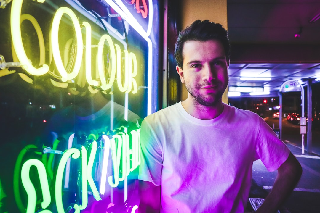 man in white crew neck t-shirt standing near purple and yellow neon signage