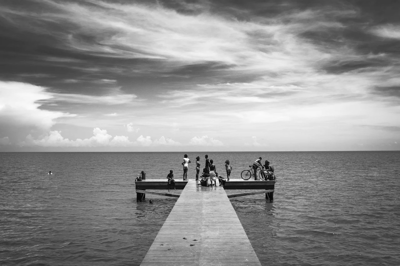 Black and white photo of a group of people at the end of a long wooden dock over the sea, under a streaked sky.