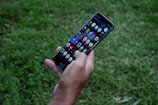 A hand holding a smartphone with a visible array of colorful app icons on the screen, surrounded by a grassy outdoor environment.
