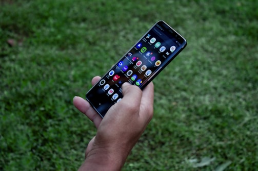 A hand holding a smartphone with a visible array of colorful app icons on the screen, surrounded by a grassy outdoor environment.