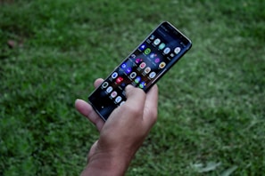 A hand holding a smartphone with a visible array of colorful app icons on the screen, surrounded by a grassy outdoor environment.