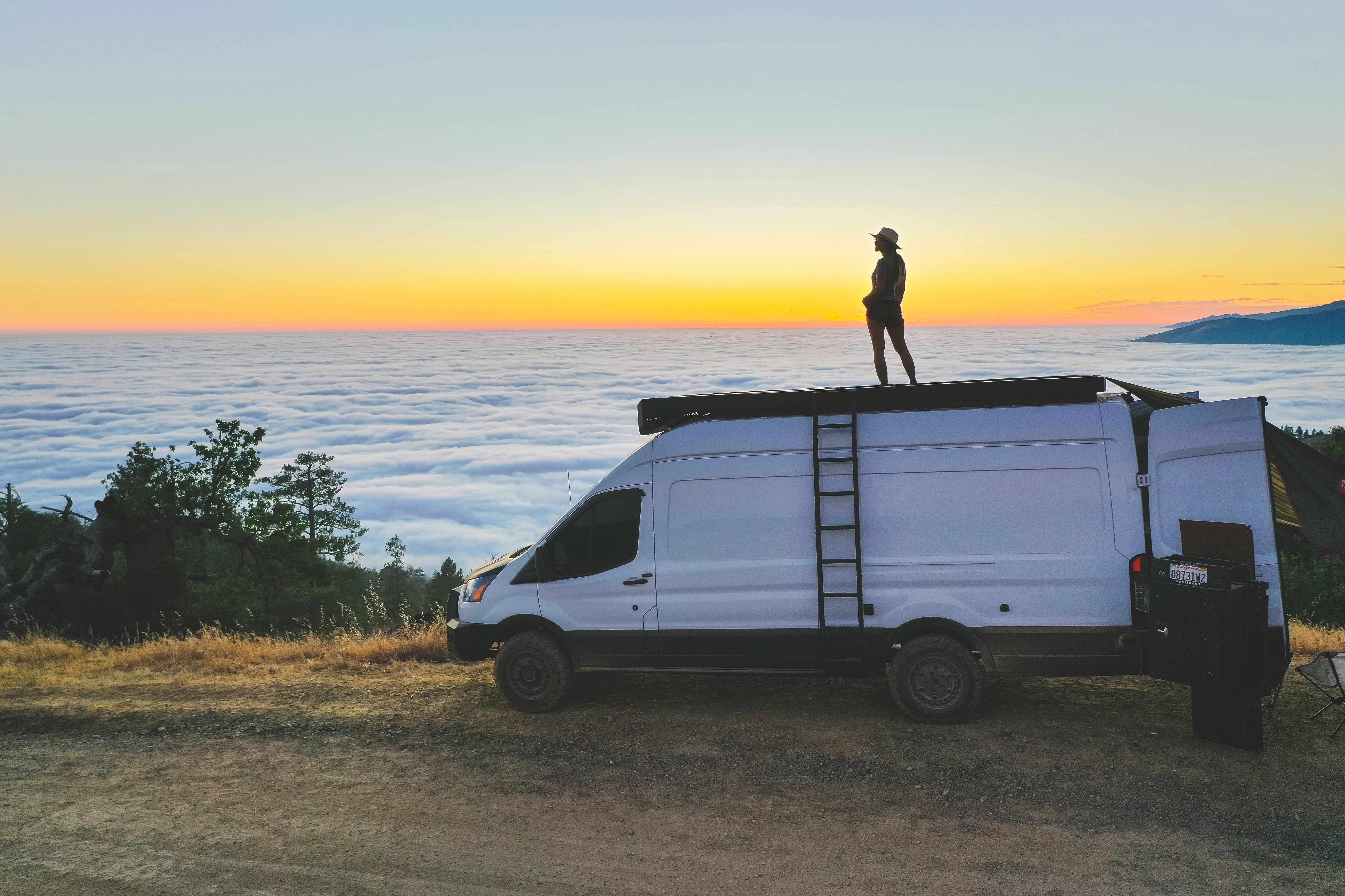 man in black jacket standing on white van near body of water during daytime