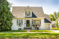 A charming suburban house with a steep roof and white walls nestled in a lush neighborhood. The front yard is well-maintained with green grass and trees. A person is tending to flower pots on the window sills, adding vibrant colors to the scene.