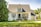 A charming suburban house with a steep roof and white walls nestled in a lush neighborhood. The front yard is well-maintained with green grass and trees. A person is tending to flower pots on the window sills, adding vibrant colors to the scene.