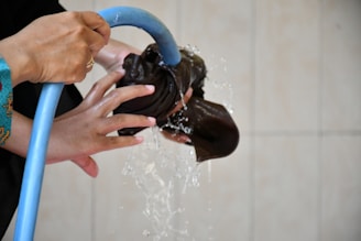 Hands holding a black cloth while being rinsed with water from a blue hose. The water is in motion, creating a dynamic scene. The background is a light, neutral color.