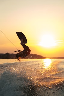 A person is wakeboarding on the water, performing an aerial trick against the backdrop of a vibrant sunset. The sun is low on the horizon, casting warm golden hues over the scene. Water droplets are splashing in the air, adding dynamic energy to the moment.