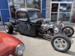 A custom-built, black, vintage hot rod truck is parked on a street. The vehicle features an exposed engine with chrome details and visible wiring. Its body has a matte finish with intricate designs on the doors. Another classic car, painted in red, is partially visible in the foreground. The scene is set in front of a storefront with glass windows reflecting the sky and surroundings.