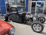 A custom-built, black, vintage hot rod truck is parked on a street. The vehicle features an exposed engine with chrome details and visible wiring. Its body has a matte finish with intricate designs on the doors. Another classic car, painted in red, is partially visible in the foreground. The scene is set in front of a storefront with glass windows reflecting the sky and surroundings.