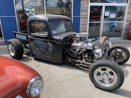 A custom-built, black, vintage hot rod truck is parked on a street. The vehicle features an exposed engine with chrome details and visible wiring. Its body has a matte finish with intricate designs on the doors. Another classic car, painted in red, is partially visible in the foreground. The scene is set in front of a storefront with glass windows reflecting the sky and surroundings.