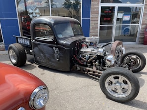 A custom-built, black, vintage hot rod truck is parked on a street. The vehicle features an exposed engine with chrome details and visible wiring. Its body has a matte finish with intricate designs on the doors. Another classic car, painted in red, is partially visible in the foreground. The scene is set in front of a storefront with glass windows reflecting the sky and surroundings.